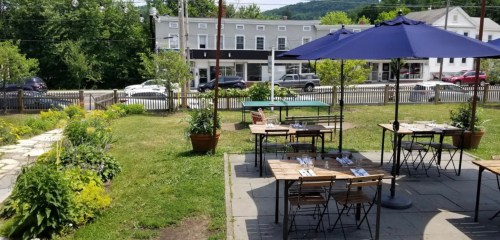 Outdoor dining area with wooden tables, umbrellas, and a view of a street lined with trees and buildings.
