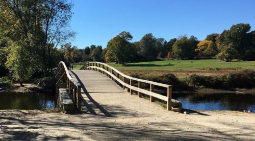 A wooden bridge spans a calm river, surrounded by trees with autumn foliage and a clear blue sky.