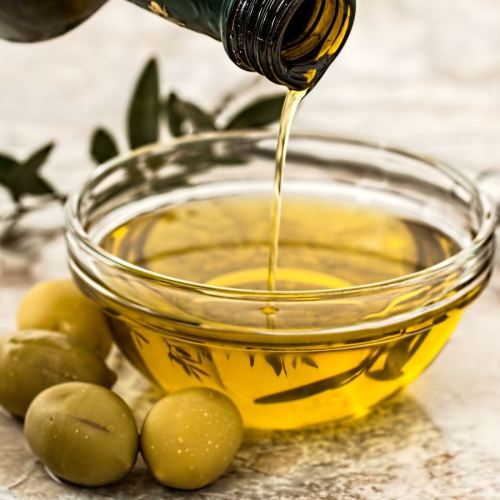 A bottle pouring olive oil into a glass bowl, surrounded by green olives and olive leaves.