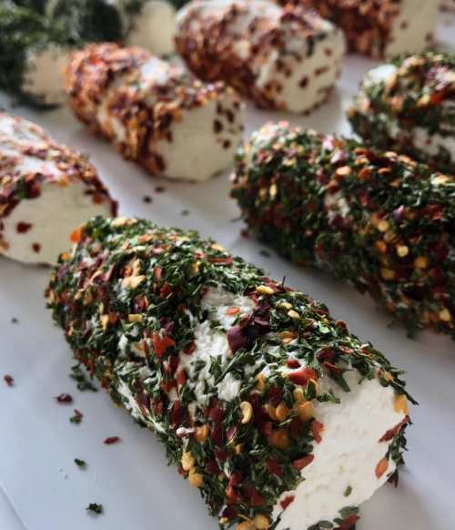 Fresh cheese logs coated in herbs and red pepper flakes, arranged on a white surface.