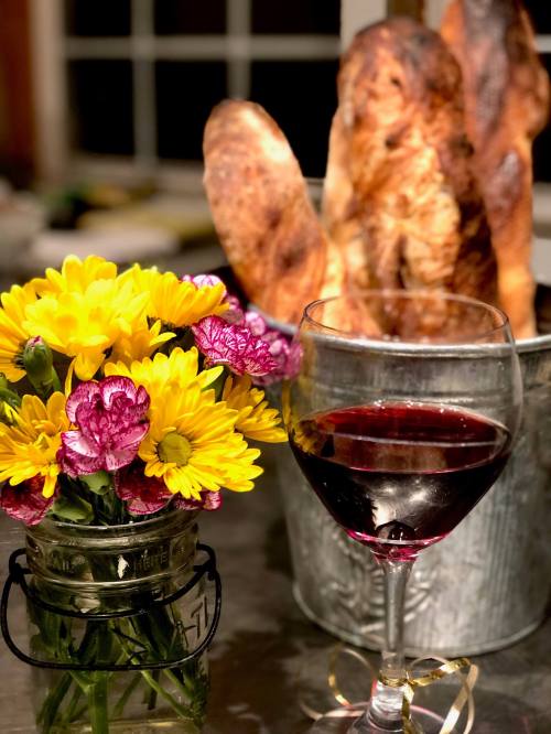 A glass of red wine beside a jar of colorful flowers and a bucket of freshly baked bread.