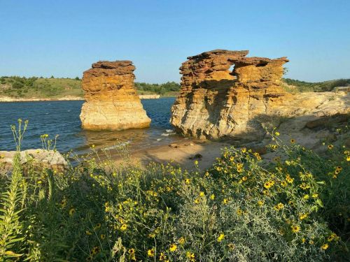 Two rock formations rise from a calm lake, surrounded by vibrant yellow wildflowers under a clear blue sky.