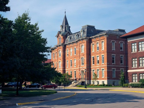 Historic brick building with a pointed tower, surrounded by trees and a paved road on a clear day.
