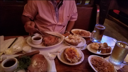 A table filled with various dishes, including meat, sides, and drinks, with a person eating in a casual setting.