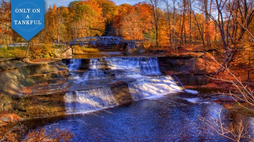 A scenic waterfall surrounded by autumn foliage, with a bridge in the background and a clear blue sky.