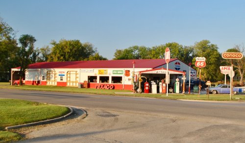 A vintage gas station with retro signage, featuring Texaco, Phillips 66, and Conoco, set in a sunny landscape.