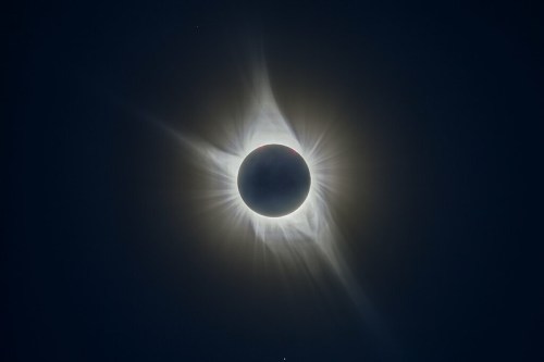 A solar eclipse with a darkened sun surrounded by radiant corona and wispy solar flares against a dark sky.