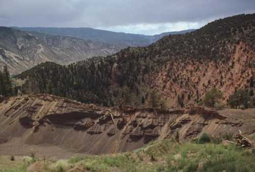 A rugged landscape featuring layered hills and mountains under a cloudy sky, with greenery in the foreground.