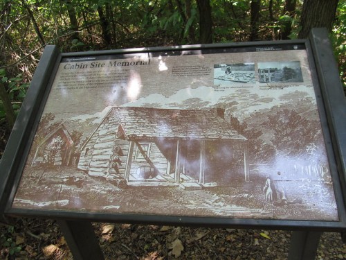 Sign depicting a cabin site memorial, featuring an illustration of a log cabin and historical information.