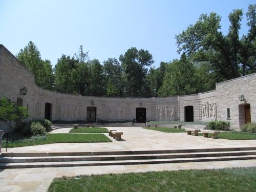 A stone courtyard surrounded by trees, featuring relief sculptures on the walls and benches on a paved area.