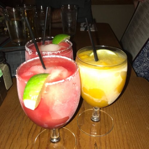 Three colorful cocktails on a wooden table, featuring a red drink with lime and a yellow drink with fruit pieces.