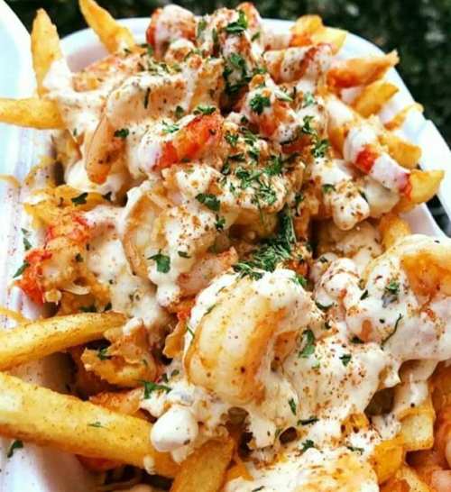 A close-up of loaded fries topped with shrimp, creamy sauce, and herbs, served in a white container.