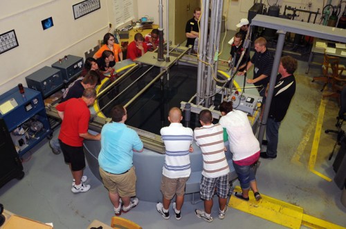 A group of students observes a demonstration around a large water tank in a lab setting.