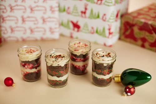 Four festive dessert jars with layers of chocolate cake and frosting, surrounded by holiday-themed gifts and ornaments.
