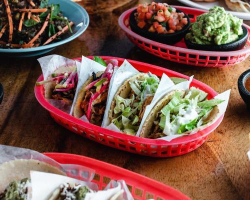 A vibrant platter of tacos in a red basket, accompanied by sides of guacamole and salsa on a wooden table.