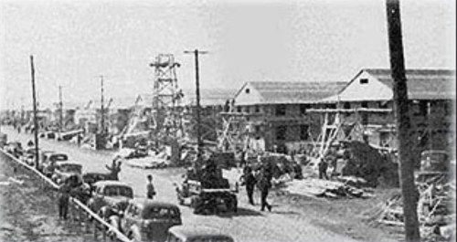 Black and white photo of a street lined with buildings and vehicles, with people and construction equipment visible.