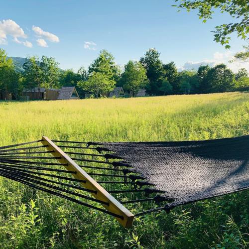 A black hammock hangs between trees in a sunny field, with distant tents and mountains in the background.
