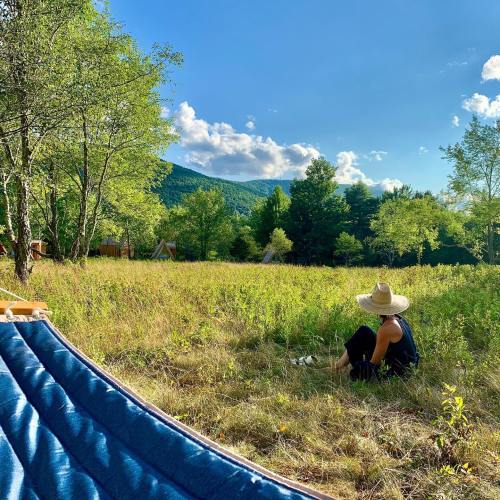 A person sits in a grassy field, wearing a straw hat, with mountains and trees in the background under a blue sky.