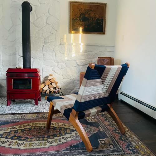 A stylish chair with a striped fabric design sits in a cozy room with a red stove and a stone wall.