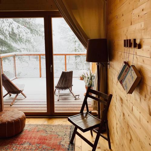 Cozy wooden interior with a chair, lamp, and books, overlooking a snowy balcony with lounge chairs.