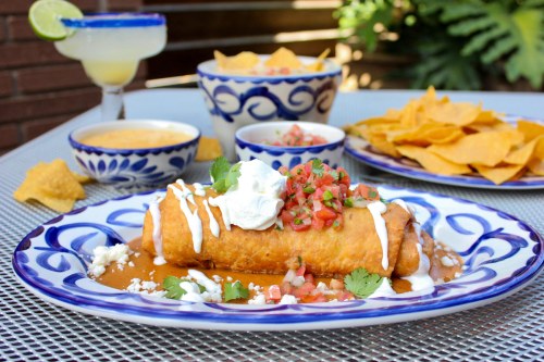 A plate of enchiladas topped with sour cream, pico de gallo, and cilantro, served with chips and a margarita.