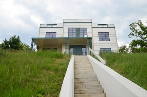 A modern two-story house on a grassy hill, featuring large windows and a staircase leading to the entrance.