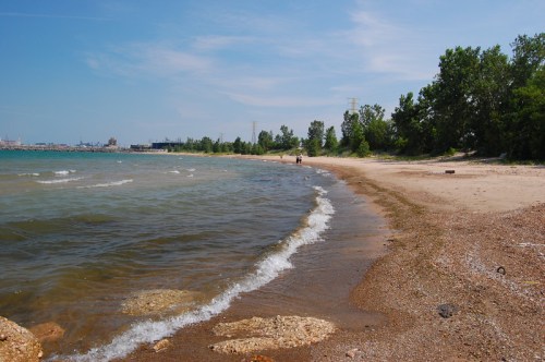 A sandy beach curves along a calm lake, with gentle waves lapping at the shore and trees lining the background.