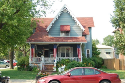 A charming blue house with a red roof, white trim, and patriotic decorations, surrounded by greenery and flowers.