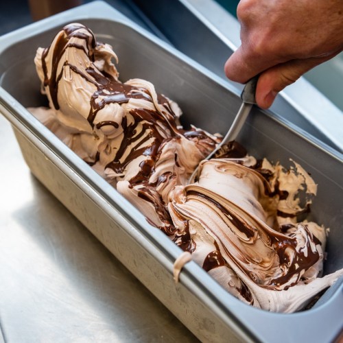 A hand scoops chocolate swirl gelato from a metal container, showcasing creamy texture and rich chocolate ribbons.