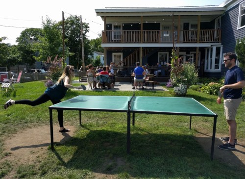A woman plays table tennis outdoors while a man watches, with people seated at a nearby patio in the background.