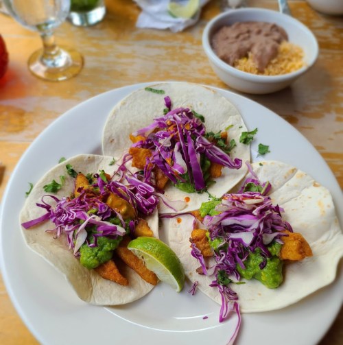 Three tacos filled with fried fish, green sauce, and purple cabbage, served with a lime wedge and a side of refried beans.