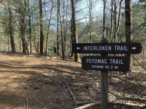 A wooden signpost points to Interloken Trail and Potomac Trail in a wooded area with a person in the background.