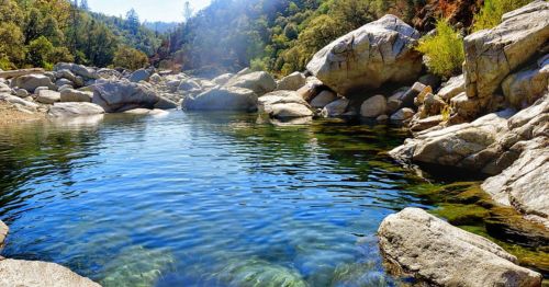 A serene river scene with clear water surrounded by large rocks and lush greenery under a bright blue sky.