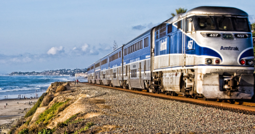 An Amtrak train travels along a coastal track with a beach and ocean in the background.