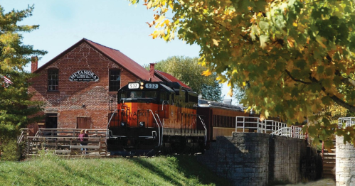 A vintage train near a historic brick building, surrounded by colorful autumn foliage.