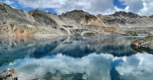 A serene mountain lake reflecting clouds and rocky terrain under a blue sky.