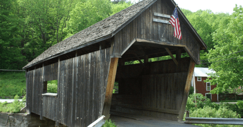 A rustic wooden covered bridge with an American flag, surrounded by lush green trees and a rural landscape.