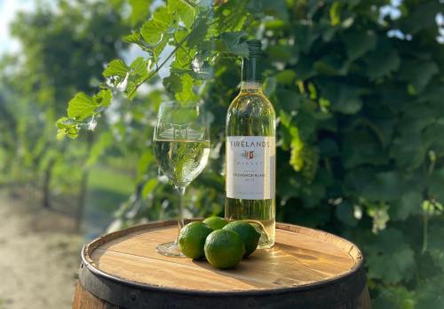 A bottle of Firelands Sauvignon Blanc and a glass of wine sit on a wooden barrel, surrounded by green vines and limes.