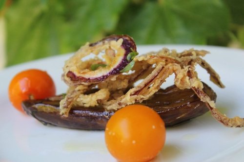 A plate featuring fried eggplant slices topped with crispy onion rings and two yellow cherry tomatoes.