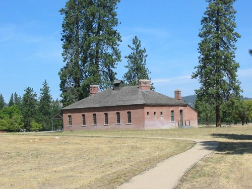 A historic brick building surrounded by tall trees and a grassy area, with a clear blue sky in the background.