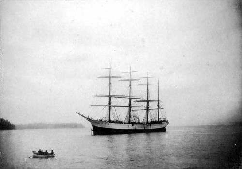 A vintage black-and-white photo of a tall ship anchored in calm waters, with a small boat nearby.