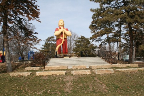 A large statue of a figure in a red robe stands on a stone platform surrounded by trees in a park.