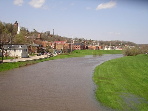A scenic view of a river alongside a grassy bank, with buildings and trees in the background under a clear sky.