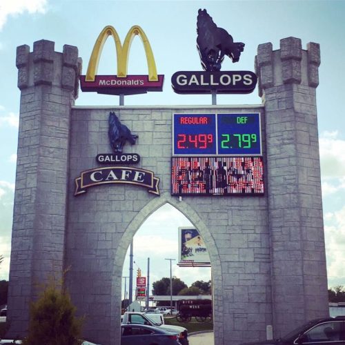 Sign for a McDonald's with a castle design, displaying gas prices and "Gallops Cafe" in front of a busy parking lot.