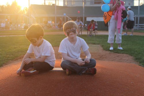 Two young boys with face paint sit on a baseball field, one on a board, during a sunny event with people in the background.