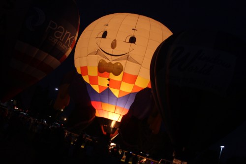 A brightly lit hot air balloon shaped like a smiling character at night, surrounded by other balloons.