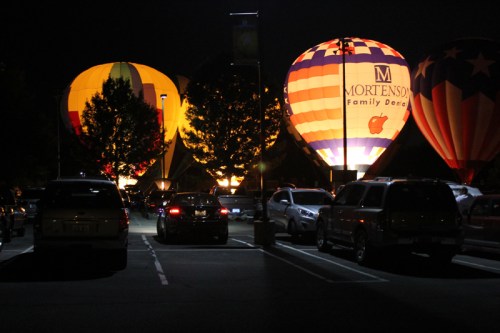 Colorful hot air balloons illuminated at night, surrounded by parked cars in a dimly lit area.