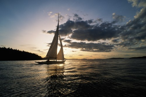A silhouette of a sailboat on calm waters at sunset, with clouds and a dark shoreline in the background.