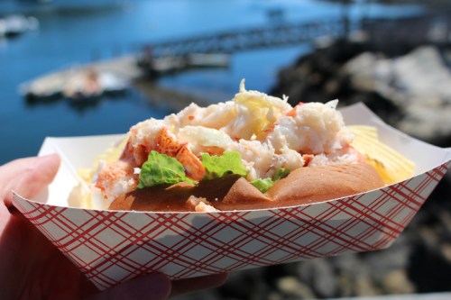 A hand holding a lobster roll with lettuce, set against a scenic waterfront view.