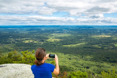 A person takes a photo of a vast green landscape from a rocky viewpoint under a cloudy sky.